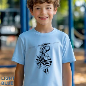 Op de afbeelding: Lichtblauw T-shirt met een zwart-witte illustratie van een stripfiguur met een staf. Het shirt heeft korte mouwen en de tekst "LIGHT BLUE" staat linksonder.