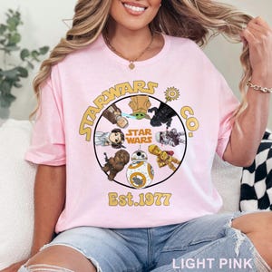 Peut inclure: T-shirt rose clair avec un motif circulaire repr&eacute;sentant des personnages de Star Wars et le texte "STAR WARS CO. Est. 1977" en or. Le t-shirt a un col rond et des manches courtes.