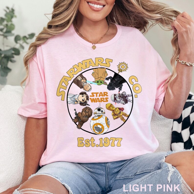 Peut inclure: T-shirt rose clair avec un motif circulaire repr&eacute;sentant des personnages de Star Wars et le texte "STAR WARS CO. Est. 1977" en or. Le t-shirt a un col rond et des manches courtes.