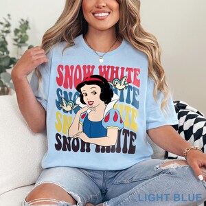 May include: Light blue t-shirt featuring a graphic of Snow White with the text "SNOW WHITE" in red, blue, yellow, and brown. The design includes two birds. The shirt is light blue.