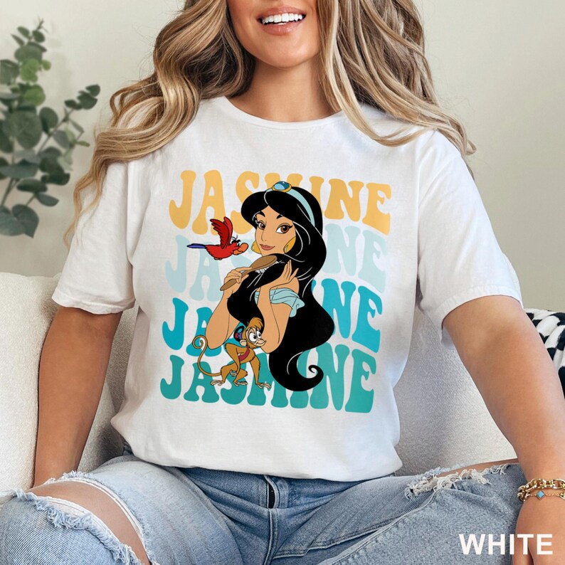 May include: White t-shirt featuring a graphic of Princess Jasmine with the word "Jasmine" in a retro font. The design includes a parrot and a monkey. The shirt is made of a soft material.