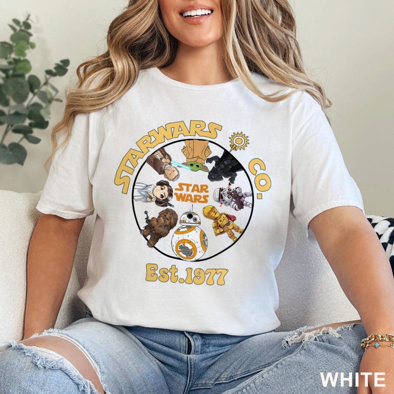 Peut inclure: T-shirt blanc avec un motif circulaire repr&eacute;sentant des personnages de Star Wars et le texte "STARWARS CO." et "Est. 1977" en jaune. Le t-shirt a des manches courtes et un col rond.