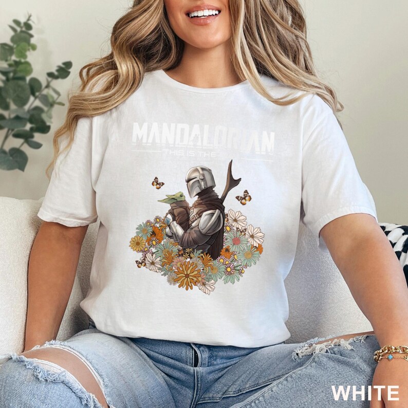 May include: White t-shirt featuring a graphic of The Mandalorian holding Grogu, surrounded by flowers and butterflies. The text "MANDALORIAN" is printed above the graphic. The shirt is made of a soft material.
