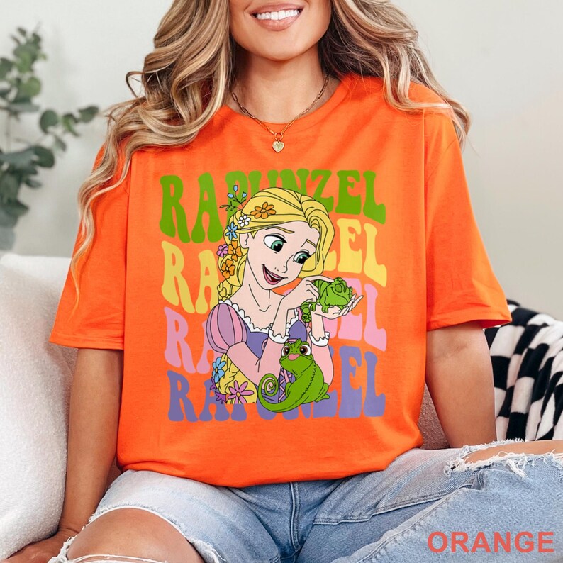 May include: Orange t-shirt featuring a graphic of Rapunzel holding a green frog, with the name "Rapunzel" in colorful, stacked text. The shirt has short sleeves and a crew neck. The word "ORANGE" is printed at the bottom.