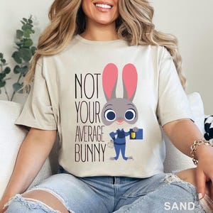 Puede incluir: Camiseta color arena con el texto "NOT YOUR AVERAGE BUNNY" y un personaje de conejo de dibujos animados. El conejo tiene orejas rosas, pelaje gris y lleva un uniforme azul y sostiene un malet&iacute;n. La palabra "SAND" est&aacute; en la parte inferior.