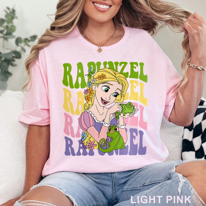 May include: Light pink t-shirt featuring a graphic of Rapunzel with the word "Rapunzel" in a retro font. The design includes a cartoon illustration of Rapunzel holding a green chameleon. The text "LIGHT PINK" is at the bottom.