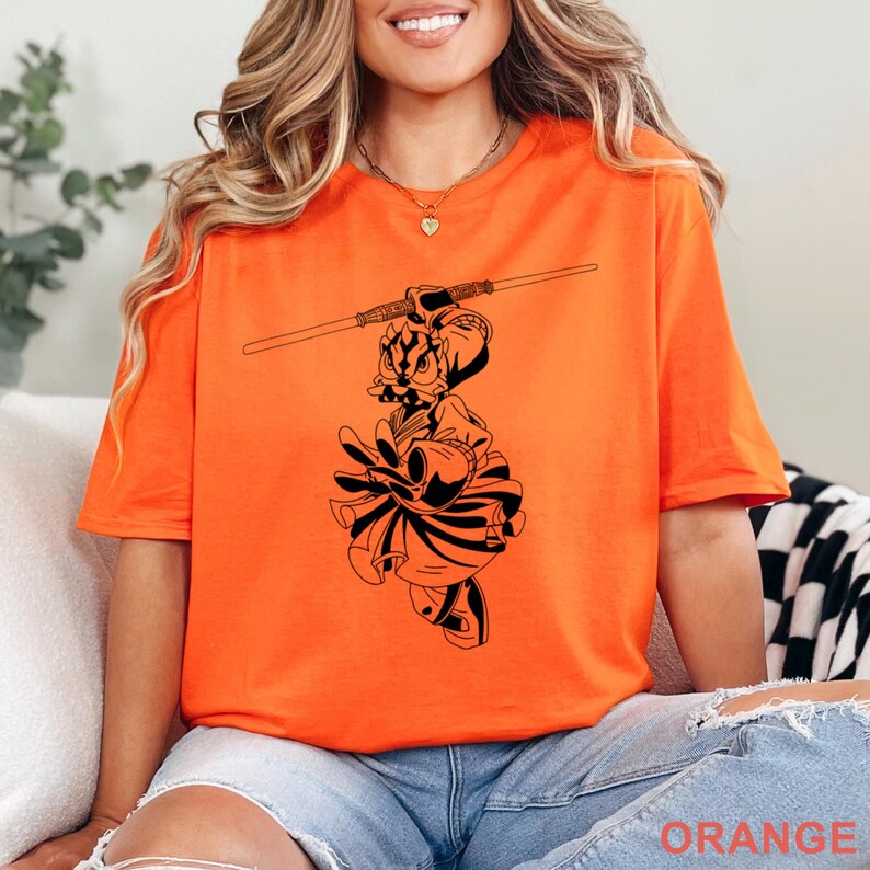 Op de afbeelding: Fel oranje t-shirt met een zwarte afbeelding van een personage met een staf. De afbeelding is een gestileerde illustratie. Het woord "ORANGE" staat onderaan het shirt. Het shirt is gemaakt van een zacht materiaal.