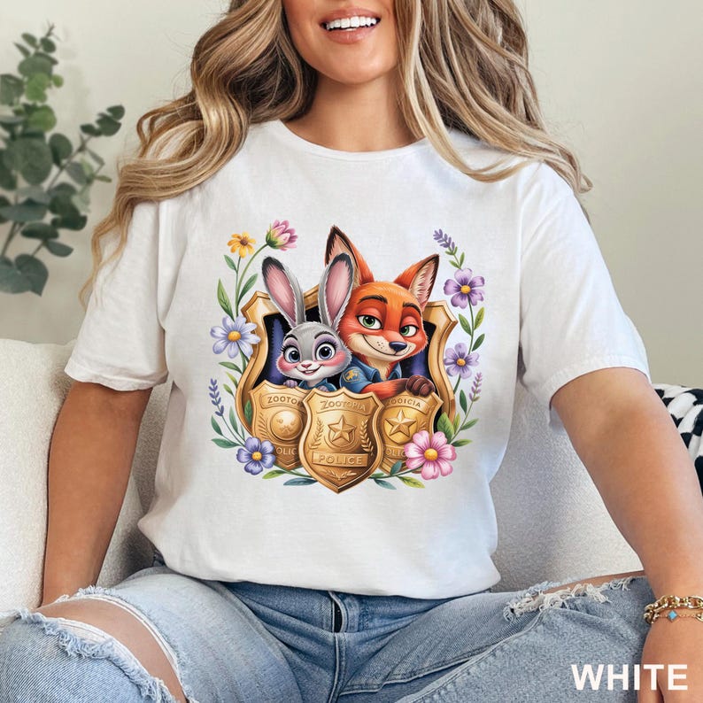 Puede incluir: Camiseta blanca con una colorida ilustraci&oacute;n de Judy Hopps y Nick Wilde de Zootopia, enmarcada por flores e insignias de polic&iacute;a. La palabra "WHITE" est&aacute; impresa en la parte inferior de la camiseta.