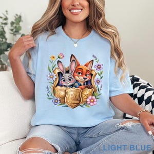 Puede incluir: Camiseta azul claro con un dibujo animado de un conejo y un zorro con insignias de polic&iacute;a, rodeados de flores. El texto "LIGHT BLUE" est&aacute; en la parte inferior. La camiseta es de cuello redondo.