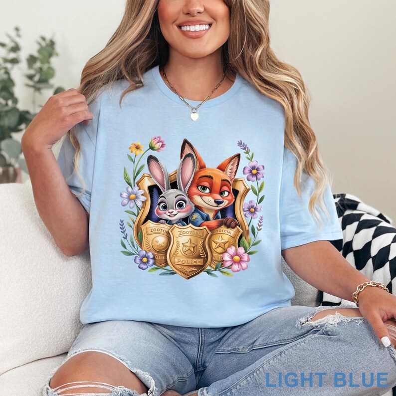 Puede incluir: Camiseta azul claro con un dibujo animado de un conejo y un zorro con insignias de polic&iacute;a, rodeados de flores. El texto "LIGHT BLUE" est&aacute; en la parte inferior. La camiseta es de cuello redondo.