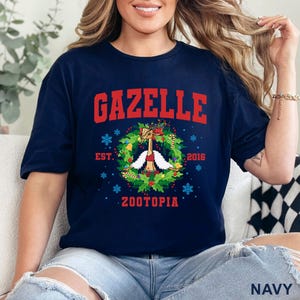 以下が含まれることがあります： 紺色のTシャツ。赤い文字で「GAZELLE」と書かれています。その下に、漫画のガゼルのキャラクターが描かれたリースデザインがあります。「EST. 2016」と「ZOOTOPIA」の文字もプリントされています。