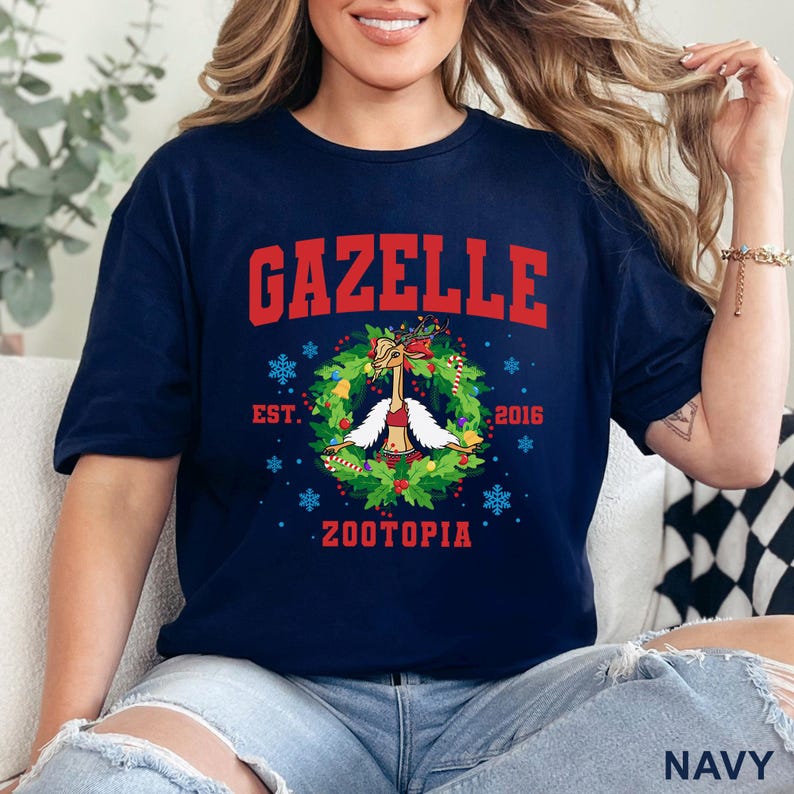 以下が含まれることがあります： 紺色のTシャツ。赤い文字で「GAZELLE」と書かれています。その下に、漫画のガゼルのキャラクターが描かれたリースデザインがあります。「EST. 2016」と「ZOOTOPIA」の文字もプリントされています。