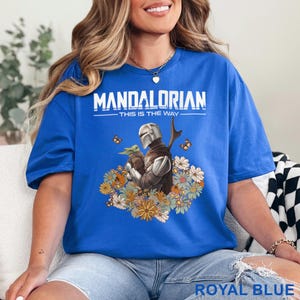 May include: Royal blue t-shirt featuring a graphic of The Mandalorian and Grogu surrounded by flowers. The text "MANDALORIAN" and "THIS IS THE WAY" are printed above the image. The shirt is a solid royal blue color.