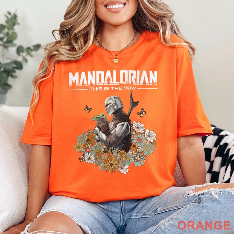 May include: Orange t-shirt featuring a graphic of The Mandalorian holding Grogu, surrounded by flowers and butterflies. The text "MANDALORIAN" and "THIS IS THE WAY" are printed above the graphic. The word "ORANGE" is printed at the bottom.