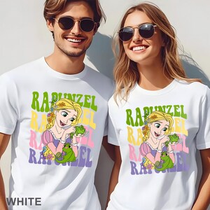 May include: White t-shirts featuring a colorful graphic of Rapunzel with the text "Rapunzel" in green, yellow, pink, and purple. The design includes an illustration of Rapunzel holding a green chameleon. The word "WHITE" is printed at the bottom of the shirt.