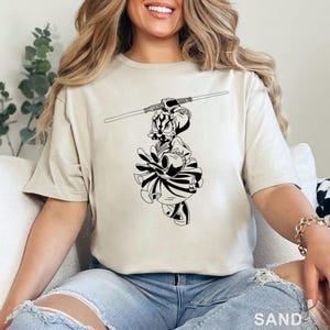 Op de afbeelding: Zandkleurig T-shirt met een zwart-witte afbeelding van een personage met een lichtzwaard. De afbeelding is een gestileerde illustratie. Het woord "SAND" staat onderaan het shirt.