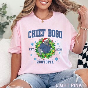 以下が含まれることがあります： 水色の文字で「CHIEF BOGO」と書かれた、薄いピンク色のTシャツ。警察の制服を着たサイの漫画のイメージが、花輪と雪の結晶に囲まれて描かれています。「ZOOTOPIA」の文字も表示されています。
