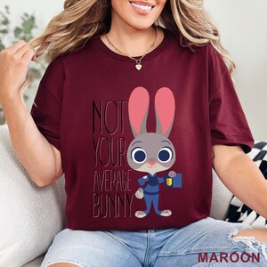 Puede incluir: Camiseta granate con un personaje de conejo de dibujos animados y el texto "NOT YOUR AVERAGE BUNNY". El conejo lleva un uniforme azul y sostiene una placa. La palabra "MAROON" est&aacute; en la parte inferior.