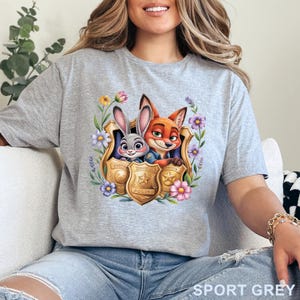 Puede incluir: Camiseta gris deportiva con un gr&aacute;fico colorido de Judy Hopps y Nick Wilde de Zootopia, enmarcado por flores y placas de polic&iacute;a. El texto "Zootopia Police" es visible en las placas. La camiseta es de material suave.
