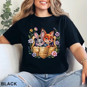 Puede incluir: Camiseta negra con un gr&aacute;fico colorido de Judy Hopps y Nick Wilde de Zootopia, enmarcado por flores y una placa de polic&iacute;a. La palabra "BLACK" est&aacute; impresa en la parte inferior de la camiseta.