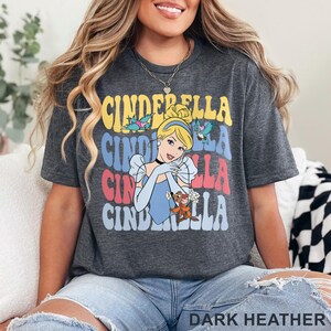 May include: Dark heather gray t-shirt featuring a colorful graphic of Cinderella with the word "CINDERELLA" stacked in multiple colors. The design includes Cinderella, birds, and her animal friends. The shirt is a comfortable, casual style.