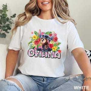 May include: White t-shirt featuring a colorful graphic of a blue alien character with a floral arrangement and the word "OHANA" in a playful font. The design includes red, yellow, and pink flowers. The shirt is a casual wear item.