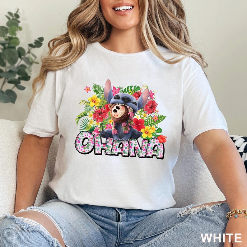 May include: White t-shirt featuring a colorful graphic of a blue alien character with a floral arrangement and the word "OHANA" in a playful font. The design includes red, yellow, and pink flowers. The shirt is a casual wear item.