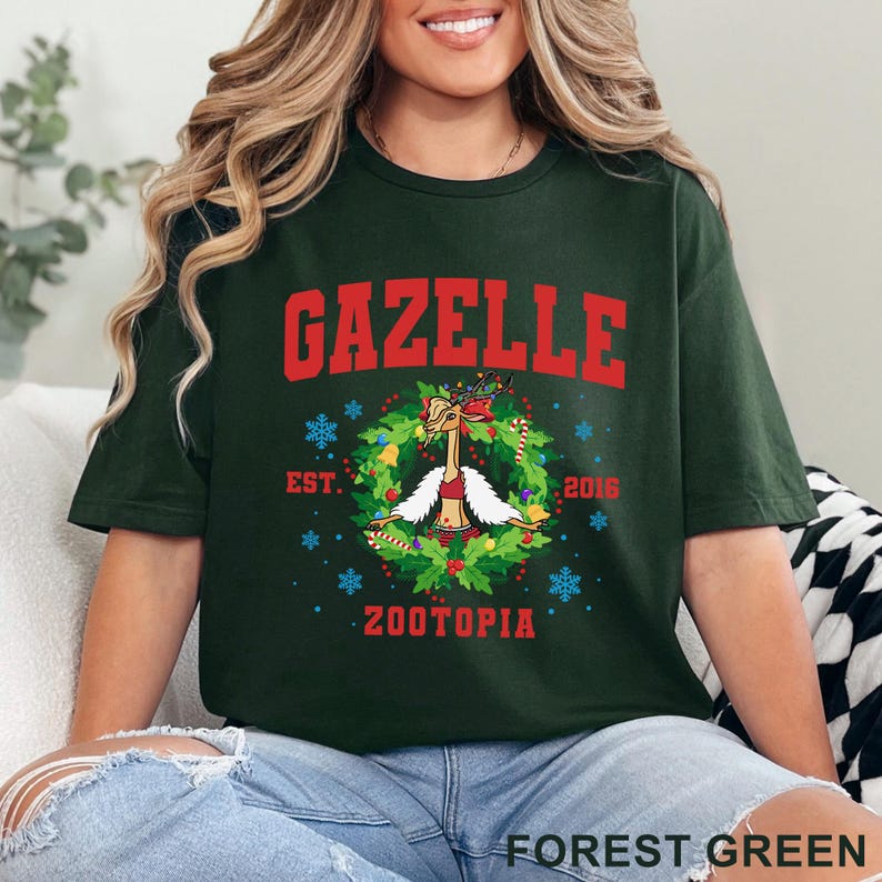 以下が含まれることがあります： 赤いブロック体の「GAZELLE」という文字が入ったフォレストグリーンのTシャツ。その下には、お祝いの衣装を着た漫画のガゼルが描かれたリースデザインがあります。「EST. 2016 ZOOTOPIA」という文字もTシャツにプリントされています。