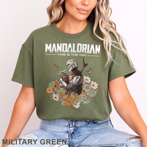 May include: Military green t-shirt featuring a graphic of The Mandalorian holding Grogu, surrounded by flowers and butterflies. The text "MANDALORIAN - THIS IS THE WAY" is printed across the top in white.