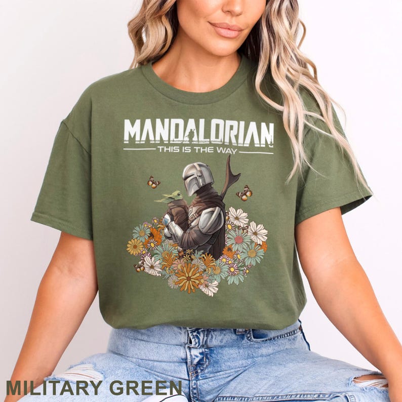 May include: Military green t-shirt featuring a graphic of The Mandalorian holding Grogu, surrounded by flowers and butterflies. The text "MANDALORIAN - THIS IS THE WAY" is printed across the top in white.