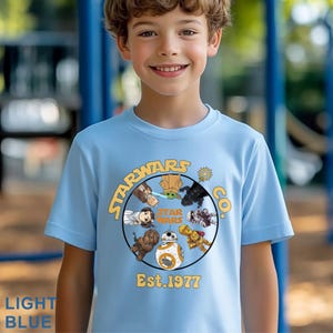 Peut inclure: T-shirt bleu clair avec un motif circulaire repr&eacute;sentant des personnages de Star Wars et le texte "STARWARS CO." et "Est. 1977." Le motif comprend Yoda, Dark Vador et BB-8. Le t-shirt est port&eacute; par un jeune.
