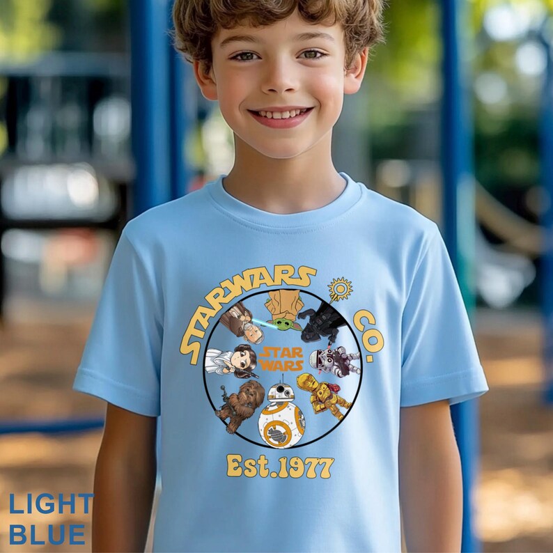 Peut inclure: T-shirt bleu clair avec un motif circulaire repr&eacute;sentant des personnages de Star Wars et le texte "STARWARS CO." et "Est. 1977." Le motif comprend Yoda, Dark Vador et BB-8. Le t-shirt est port&eacute; par un jeune.