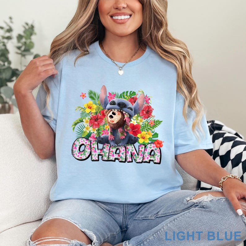May include: Light blue t-shirt featuring a colorful graphic of a cartoon character with a floral arrangement and the word "OHANA". The graphic includes a blue character wearing a hat, surrounded by red, yellow, and pink flowers. The text "LIGHT BLUE" is at the bottom.