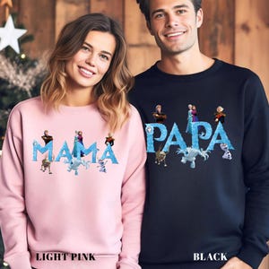 Op de afbeelding: Lichtroze en zwarte sweatshirts met de woorden "MAMA" en "PAPA" in blauw, met personages uit een populaire animatiefilm. Het lichtroze sweatshirt heeft de tekst "LIGHT PINK" onderaan. Het zwarte sweatshirt heeft de tekst "BLACK" onderaan.