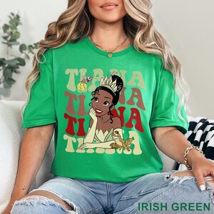 May include: Irish green t-shirt featuring a graphic of Princess Tiana with the text "Tiana" in a retro font. The design includes a frog and a firefly. The shirt is a short-sleeved crew neck.