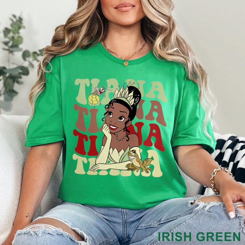 May include: Irish green t-shirt featuring a graphic of Princess Tiana with the text "Tiana" in a retro font. The design includes a frog and a firefly. The shirt is a short-sleeved crew neck.