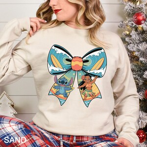 Puede incluir: Una sudadera de color arena con un gran dise&ntilde;o de lazo con Stitch y Lilo de Lilo & Stitch. El lazo tiene un patr&oacute;n de olas turquesas y blancas con tablas de surf amarillas. Los personajes est&aacute;n en una escena de playa. La palabra "SAND" est&aacute; impresa en la parte inferior.
