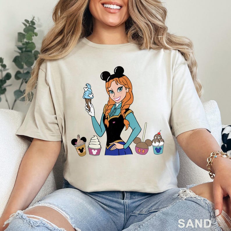May include: Sand-colored t-shirt featuring a cartoon illustration of a character with red hair and a blue ice cream cone. The character is surrounded by ice cream treats. The word "SAND" is printed at the bottom.