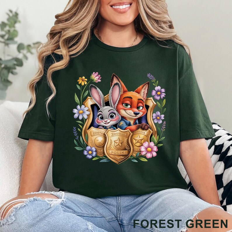 Puede incluir: Camiseta verde bosque con una ilustraci&oacute;n de dibujos animados de un conejo y un zorro dentro de una placa de polic&iacute;a dorada. La camiseta tiene el texto "FOREST GREEN" en la parte inferior. El dise&ntilde;o est&aacute; rodeado de flores coloridas.