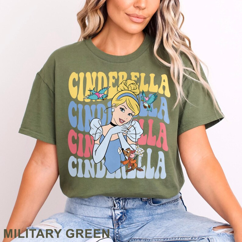 May include: Military green t-shirt featuring a colorful graphic of Cinderella with the text "Cinderella" in a retro font. The design includes Cinderella, birds, and her animal friends. The shirt is a comfortable, casual style.