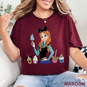 May include: Maroon t-shirt featuring a cartoon illustration of a character holding an ice cream cone, with other ice cream treats. The character has red hair and is wearing a black top and blue skirt. The text "MAROON" is at the bottom.