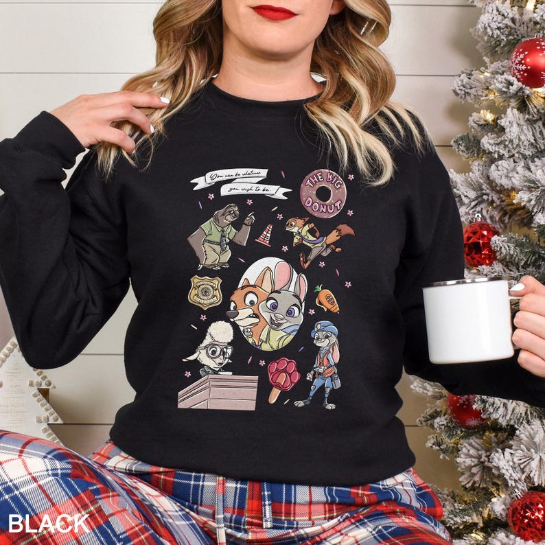Pu&ograve; includere: Felpa girocollo nera con personaggi dei cartoni animati del film "Zootropolis". Il design include immagini di un bradipo, una volpe, un coniglio e altri personaggi, insieme alle parole "The Big Donut" e "You can be anything you wish to be."