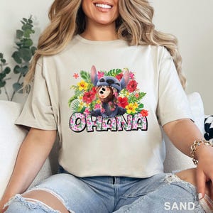 May include: Sand-colored t-shirt featuring a colorful graphic of a blue alien character with a floral design and the word "OHANA" in a playful font. The design includes tropical flowers and foliage.