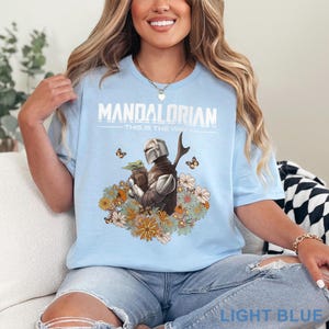 May include: Light blue t-shirt with the text "MANDALORIAN" and "THIS IS THE WAY". The graphic features the Mandalorian and Grogu surrounded by flowers and butterflies. The shirt is light blue.