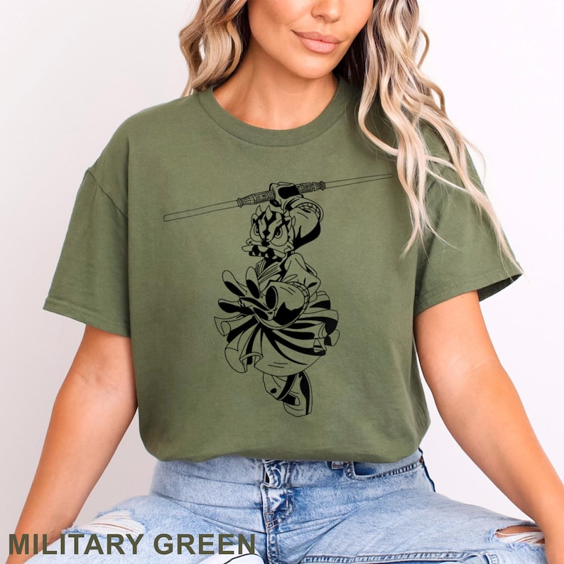 May include: Military green t-shirt featuring a black graphic of a cartoon character holding a lightsaber. The character is dressed in a warrior outfit. The text "MILITARY GREEN" is at the bottom of the image.