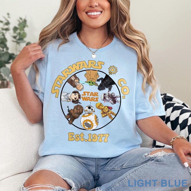 May include: Light blue Star Wars Co. t-shirt featuring a circular design with various Star Wars characters, including BB-8, Chewbacca, and Yoda. The design also includes the words 'Star Wars' and 'Est. 1977' in yellow lettering. The shirt is made of a soft material.