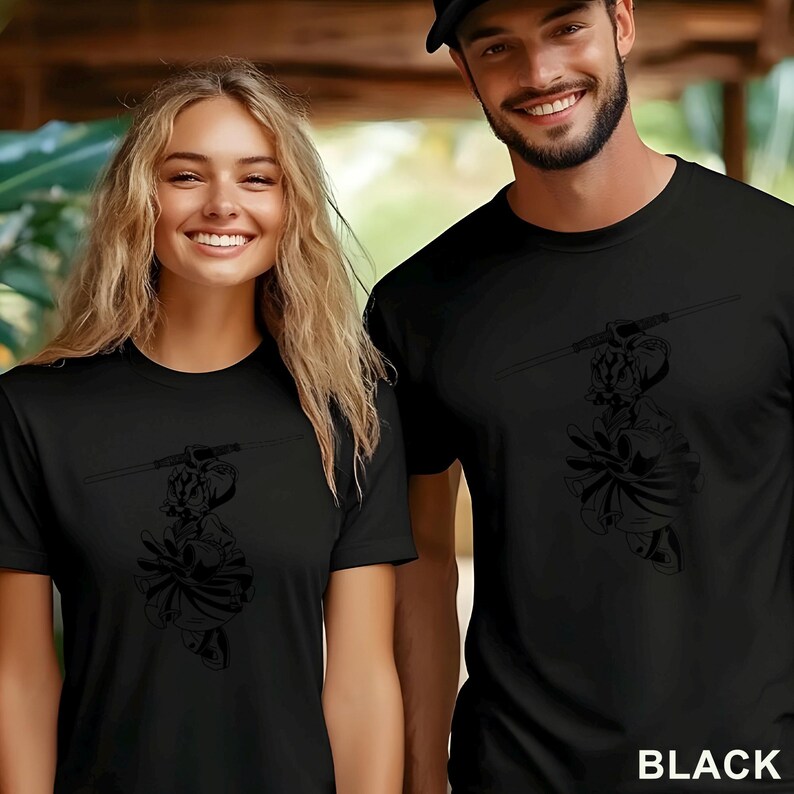 Op de afbeelding: Zwarte t-shirts met een grafisch ontwerp van een figuur met een zwaard. Het woord "BLACK" staat in het wit rechtsonder. Twee mensen dragen de shirts.