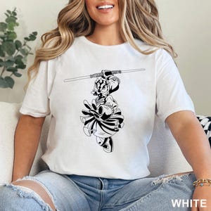Op de afbeelding: Wit t-shirt met een zwart-wit illustratie van een personage met een lichtzwaard. De afbeelding is gecentreerd. Het woord "WHITE" staat in het wit rechtsonder op het t-shirt.