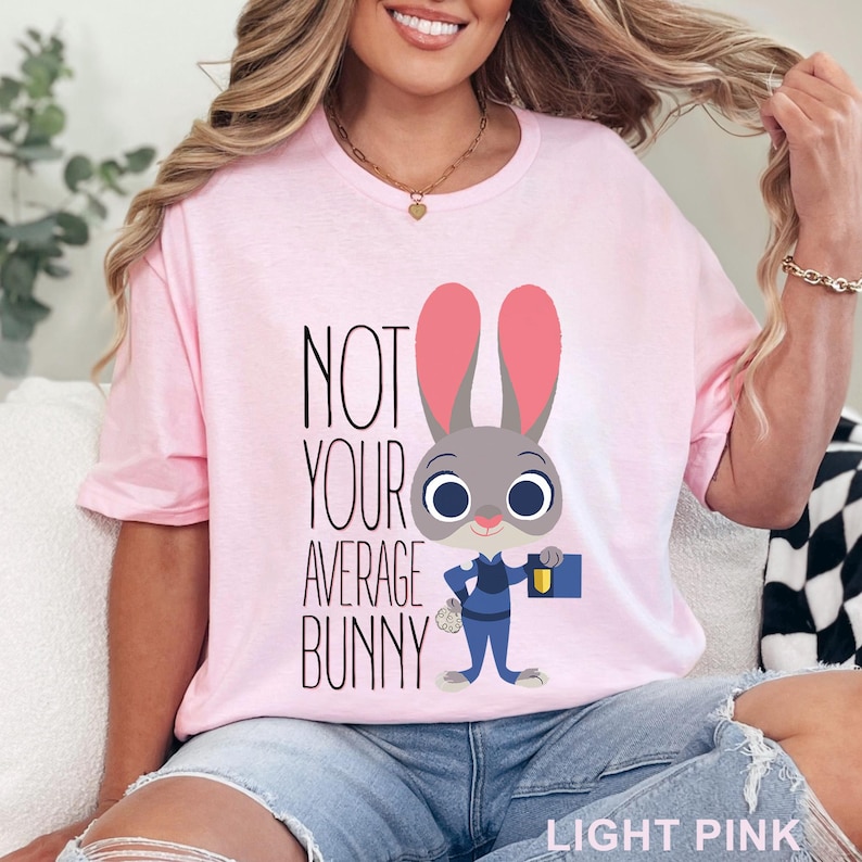 May include: Light pink t-shirt with the text "NOT YOUR AVERAGE BUNNY" and a cartoon bunny character wearing a blue uniform and holding a briefcase. The shirt is a crew neck style.