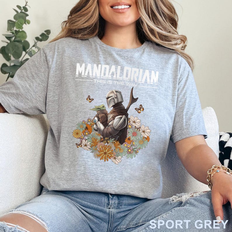 May include: Sport grey t-shirt featuring a graphic of the Mandalorian and Grogu surrounded by flowers and butterflies. The text "MANDALORIAN" and "THIS IS THE WAY" are printed above the graphic.